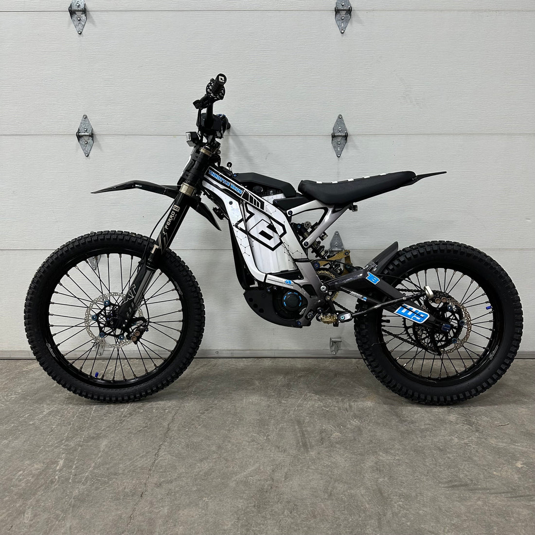 Sur-Ron Light Bee X electric motorcycle with white and black custom graphics kit, featuring geometric frame decals, fork guards, and battery housing wraps, displayed in a garage setting